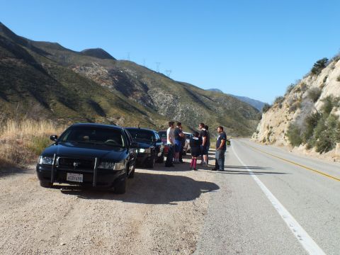 2003 Ford Crown Victoria - Dark full-size sedan parked at a roadside turnout, several people standing and talking nearby