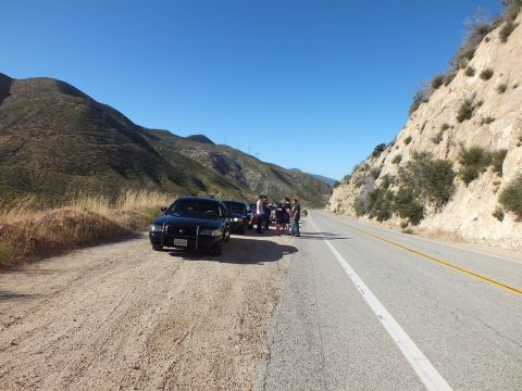 2003 Ford Crown Victoria - Dark sedan parked on a dirt shoulder with people gathered, mountain road curving past a rock face