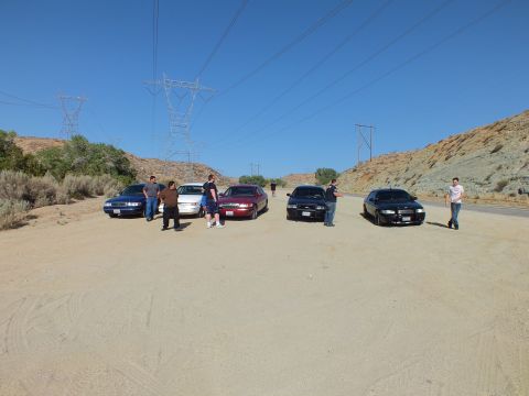 2003 Ford Crown Victoria - Five sedans lined up facing camera in a sandy lot, owners standing alongside under transmission towers