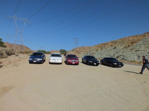 2003 Ford Crown Victoria - Lineup of full-size sedans in a desert clearing beneath steel transmission towers, hillside behind