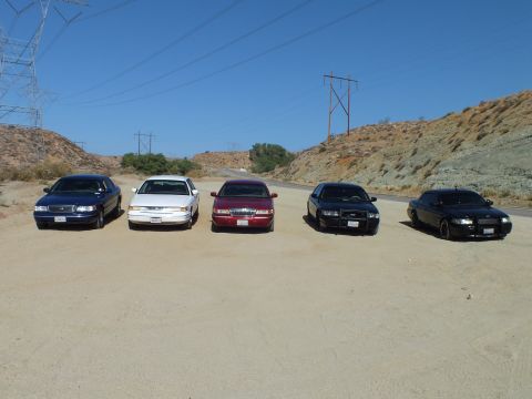 2003 Ford Crown Victoria - Full-size sedans parked in a row in a desert clearing, transmission towers and hillside behind