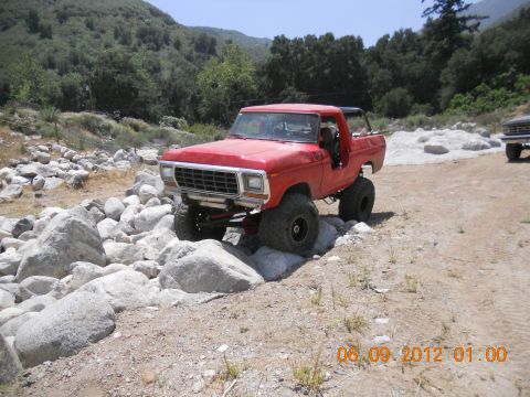1986 Ford Bronco - This lifted red Ford Bronco is shown off-roading over rocks. Another vehicle is visible in the background.