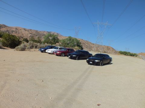 2003 Ford Crown Victoria - Row of sedans parked across a sandy clearing under power lines and desert hillside