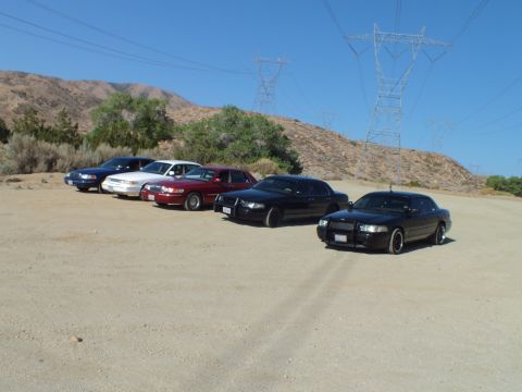 2003 Ford Crown Victoria - Lineup of sedans in a dirt clearing led by a black car, transmission towers on the hill behind
