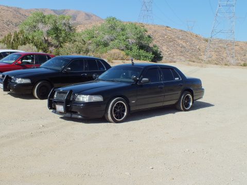 2003 Ford Crown Victoria - Blacked-out full-size sedan with push bar and dark rims beside other cars under power lines