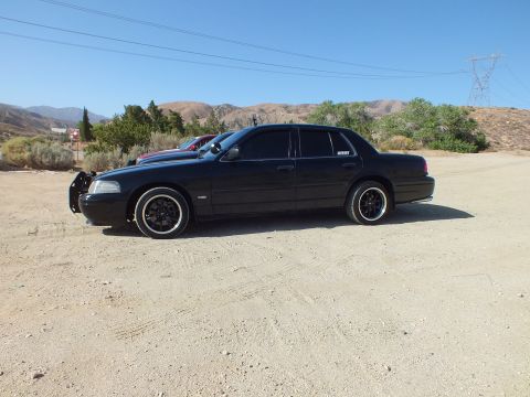 2003 Ford Crown Victoria - Profile of a blacked-out full-size sedan with push bar on dark rims, transmission towers behind