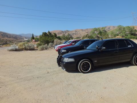 2003 Ford Crown Victoria - Lowered black full-size sedan sits on a dirt lot beside other cars under a transmission tower and dry hills