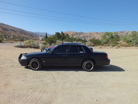 2003 Ford Crown Victoria - Black four-door sedan shown in side view on packed dirt with mountains and blue sky behind