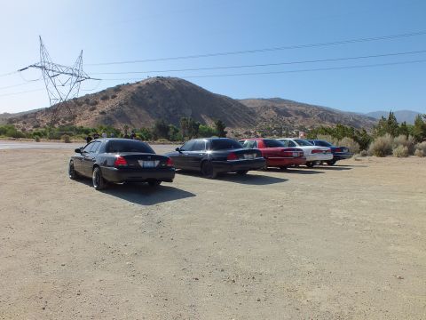 2003 Ford Crown Victoria - Dark and maroon sedans angle-parked on a dirt lot under a tall power pylon and brown hill