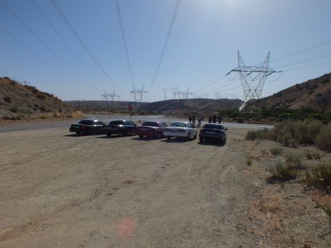 2003 Ford Crown Victoria - cluster of sedans and people gather on a dirt pullout next to power pylons and dry hills