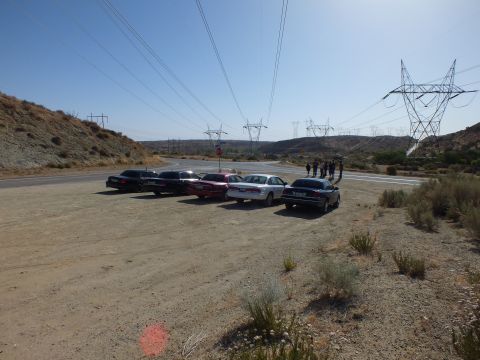 2003 Ford Crown Victoria - Several sedans angle-park on a dirt lot as people stand by the roadside under power pylons