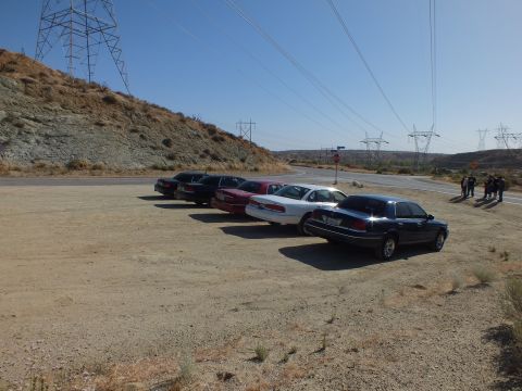 2003 Ford Crown Victoria - line of sedans sits on a dirt lot beside a hill cut as people stand near the road and pylons