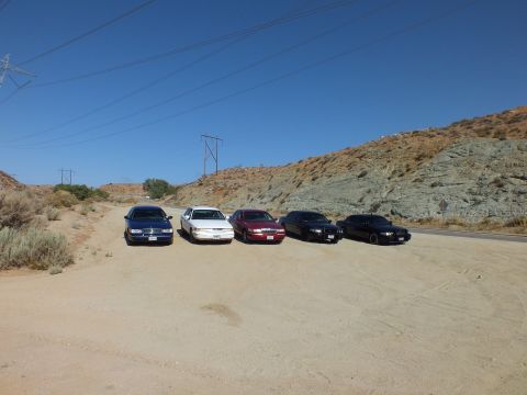 2003 Ford Crown Victoria - Blue, white, maroon and black sedans line up nose-out on sandy ground near a transmission tower