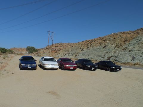 2003 Ford Crown Victoria - Five sedans sit nose-out on a dirt lot against a layered rock hillside under clear sky