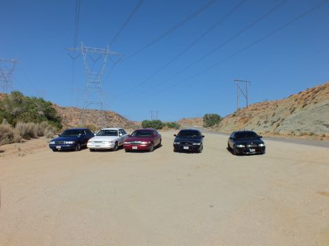 2003 Ford Crown Victoria - Blue, white, maroon and black sedans line up on sand as a road runs between rocky hillsides