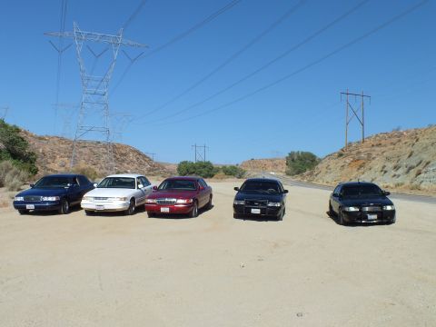 2003 Ford Crown Victoria - Mixed-color sedan lineup faces the camera on a dirt lot beneath tall steel power pylons