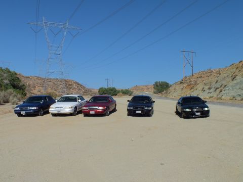 2003 Ford Crown Victoria - Five Panther-platform sedans line up facing forward on a dirt lot under power lines and hills