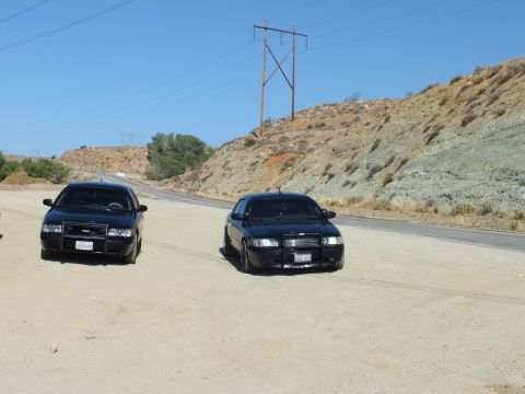 2003 Ford Crown Victoria - Pair of black Ford-style sedans sit on a desert shoulder below a power-line tower, hills behind.
