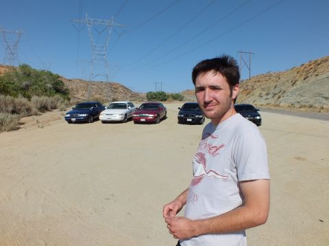 2003 Ford Crown Victoria - Five parked sedans, including a maroon and a white car, line up behind a man on a desert lot.