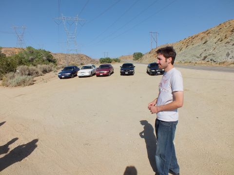2003 Ford Crown Victoria - Row of five sedans parked on dirt as a man looks on, power towers across the hillside.