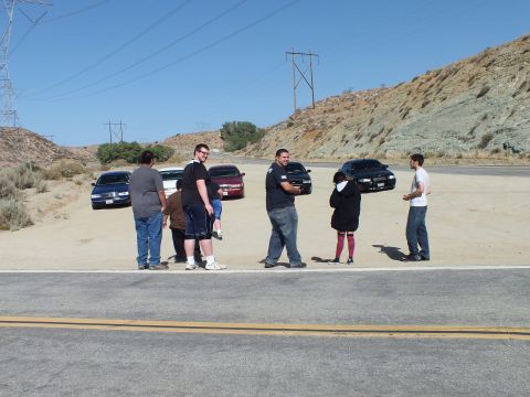 2003 Ford Crown Victoria - People mingle on a desert lot; three sedans and transmission towers stand in the background.