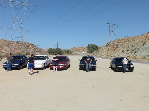 2003 Ford Crown Victoria - Five owners stand at the fronts of their parked sedans on a sandy pullout, towers behind.