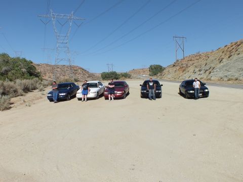 2003 Ford Crown Victoria - Group lines up before five sedans on a sandy lot under tall transmission towers.