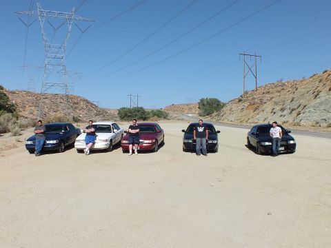 2003 Ford Crown Victoria - Each person stands by a sedan front on a sandy pullout, power lines crossing the sky.