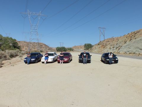 2003 Ford Crown Victoria - Owners pose at their sedans on a dirt pullout below desert hills and power towers.