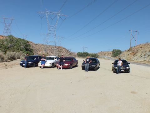 2003 Ford Crown Victoria - People gather around five parked sedans on a sandy pullout with power lines overhead.
