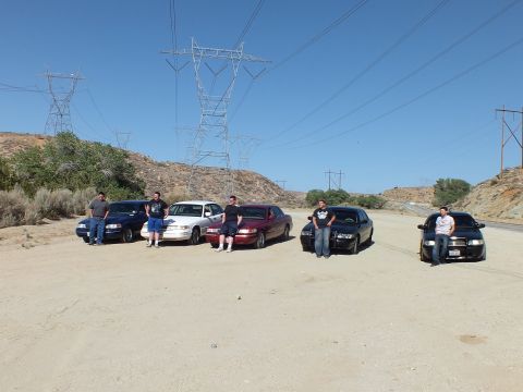 2003 Ford Crown Victoria - Owners line up at the fronts of five parked sedans under tall transmission towers.