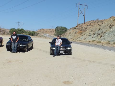 2003 Ford Crown Victoria - Two men sit on the hoods of black sedans at a sandy roadside under a power-line tower.