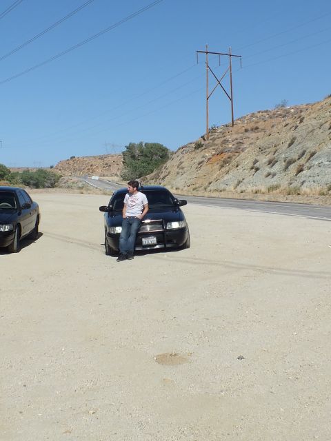 2003 Ford Crown Victoria - Man sits on the hood of a black sedan on a sandy lot, hills and power tower behind.