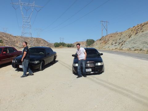 2003 Ford Crown Victoria - pair of blacked-out sedans with a man leaning on the front fender in a desert pullout