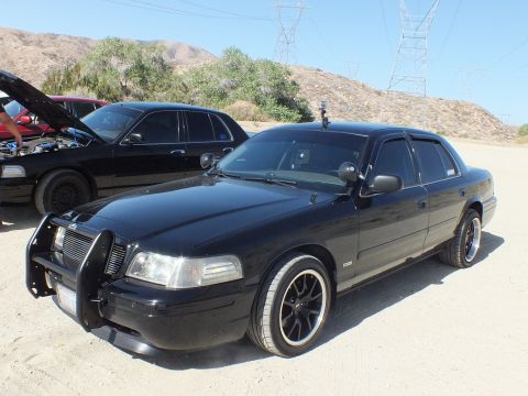 2003 Ford Crown Victoria - Two black full-size sedans parked side by side under transmission towers