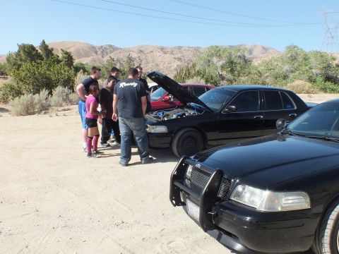 2003 Ford Crown Victoria - Group inspecting the engine bay of a black sedan with hood up at the desert meet