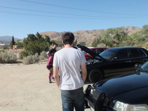 2003 Ford Crown Victoria - Onlookers gathered at the raised hood of a black sedan on a dirt lot