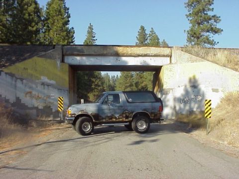 1989 Ford Bronco - A lifted full-size Ford Bronco with a removable hardtop sits broadside under a concrete railroad underpass on a cracked asphalt road, frame…