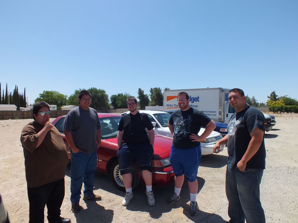 Five men posing in front of red Mercury Grand Marquis sedan in gravel lot