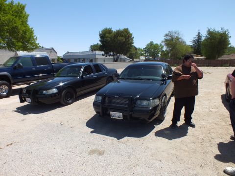 2003 Ford Crown Victoria - Pair of blacked-out Crown Victoria sedans and a dark pickup on a dirt lot, man at right with phone