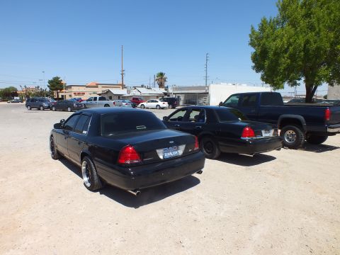 2003 Ford Crown Victoria - Rear three-quarter view of two black Crown Victorias and a pickup, town buildings behind