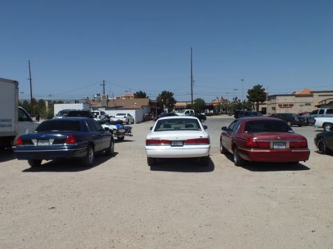 2003 Ford Crown Victoria - Three full-size sedans rear-first on dirt with shops and power lines in background