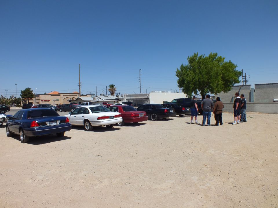 Row of parked full-size sedans with group of people gathered at right in dirt lot