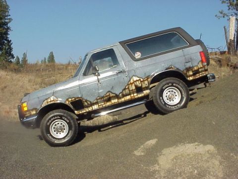 1989 Ford Bronco - Driver-side profile of a lifted gray full-size two-door Ford Bronco flexed with its rear wheels up on a dirt mound, showing a black removab…