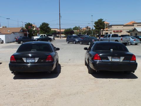 2003 Ford Crown Victoria - Rear view of two black Crown Victoria sedans with dual exhaust on a dirt lot