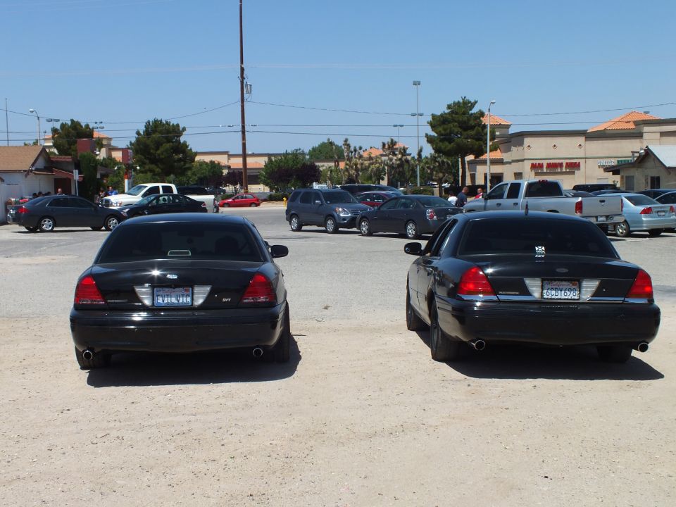 Two black full-size sedans with dual exhaust parked rear-on in dirt lot