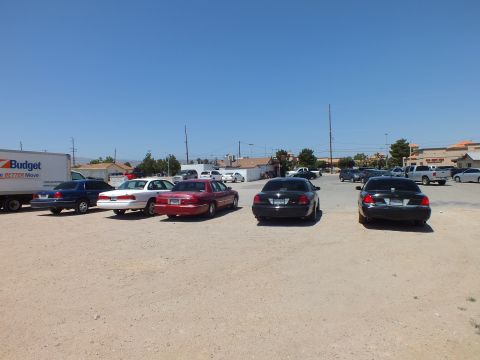 2003 Ford Crown Victoria - Mixed sedans and a dark BMW lined up on dirt near a Budget box truck and shops