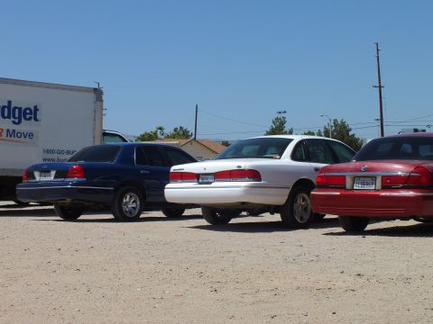 2003 Ford Crown Victoria - Close rear view of blue, white and red full-size sedans next to a Budget box truck