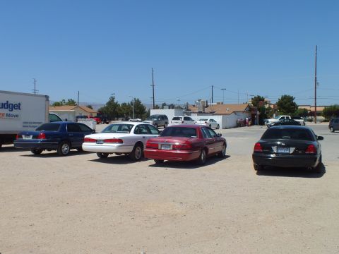2003 Ford Crown Victoria - Blue, white, maroon and black sedans parked in a dusty lot with a Budget truck and homes behind