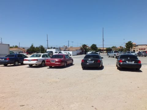 2003 Ford Crown Victoria - Blue, white, maroon and black sedans on a dusty lot, low commercial buildings across the street